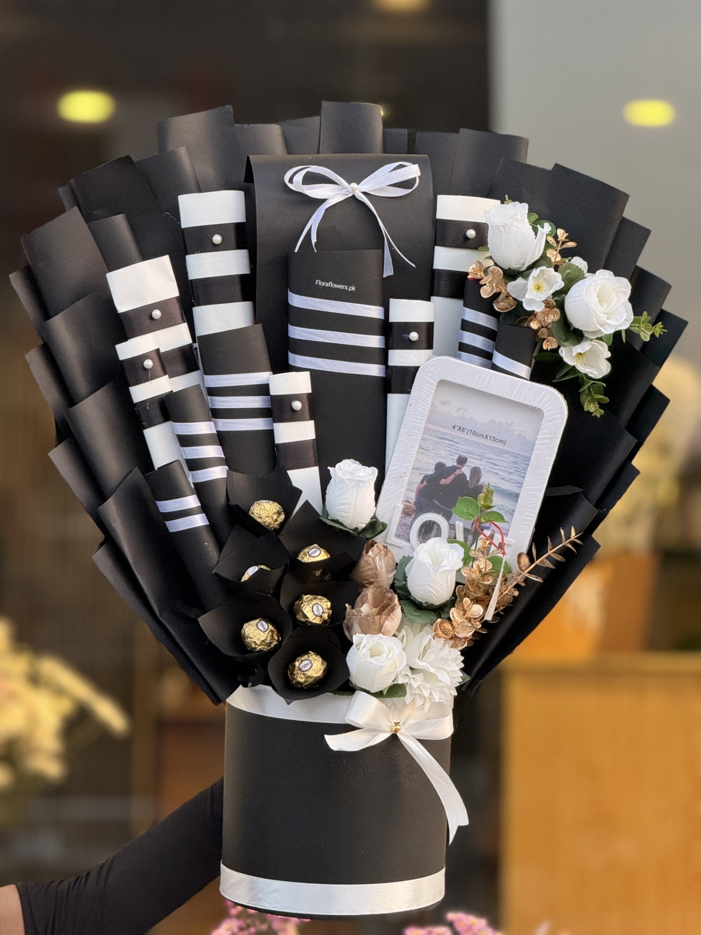 A gift arrangement featuring a photo frame, chocolate bars, and decorative elements in black, white, and gold colors.