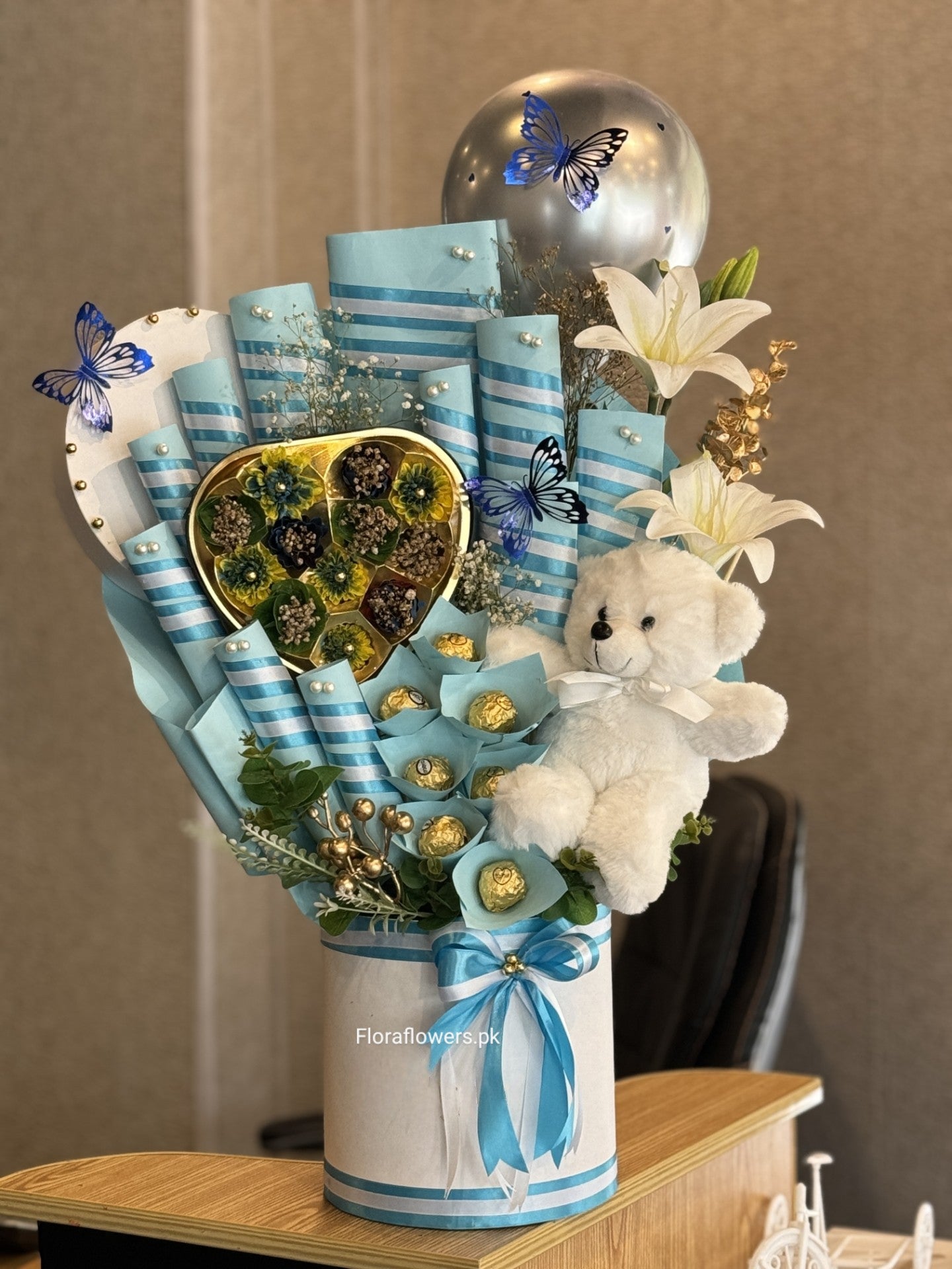 A candy bouquet arrangement featuring an assortment of chocolates including Snickers, Mars, KitKat, Dairy Milk, and Ferrero Rocher, adorned with blue ribbons, artificial blue flowers, and a small white teddy bear.
