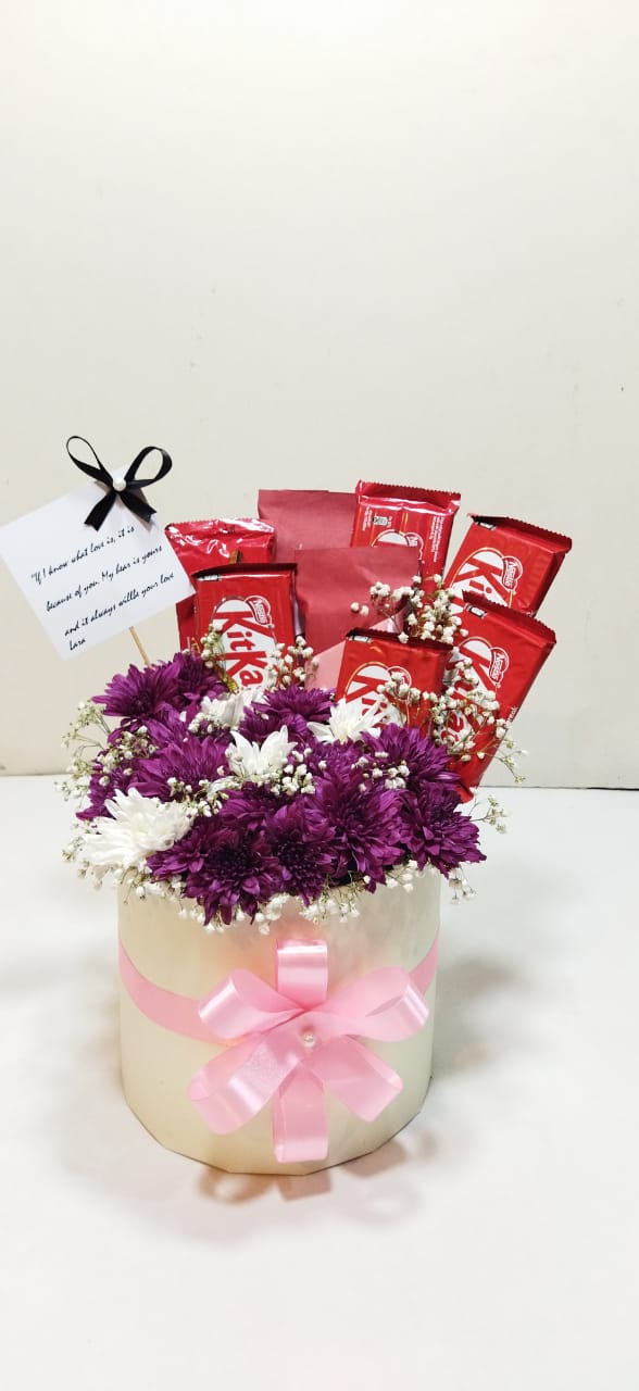 A gift basket containing fresh white and purple chrysanthemums, baby's breath, and an assortment of chocolates, with a customized tag attached.