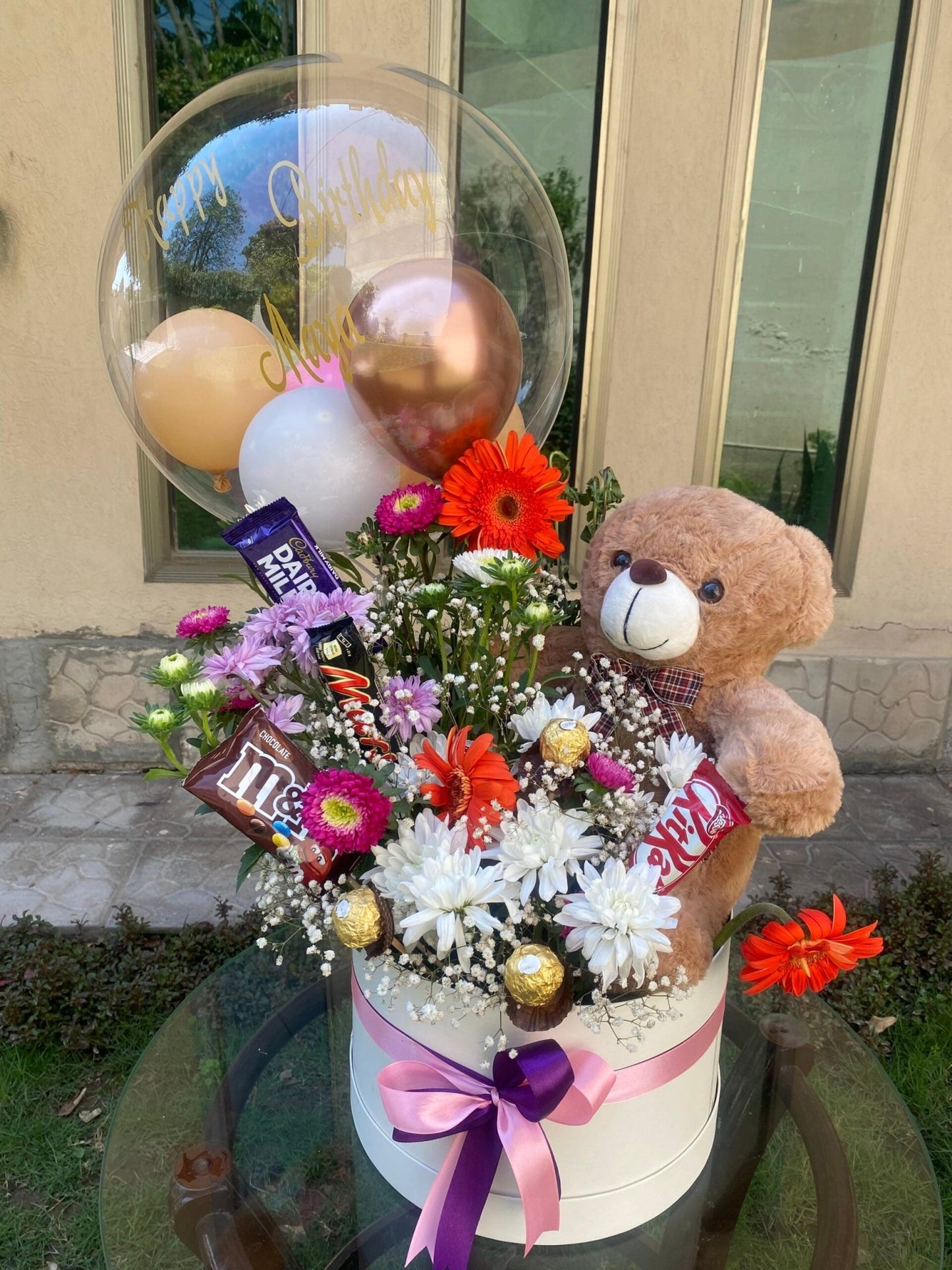 A gift basket arrangement featuring a teddy bear, various chocolates including Ferraro Rocher and M&Ms, a transparent balloon, and an assortment of flowers such as Chrysanthemums and Gerbera. The arrangement is presented in a white container with a pink ribbon.