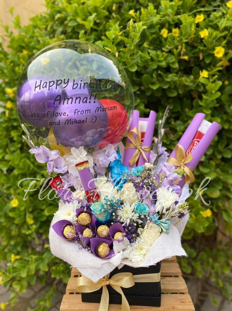 A gift basket arrangement featuring a balloon with customizable text, an assortment of colorful flowers including roses and chrysanthemums, and chocolate candies.