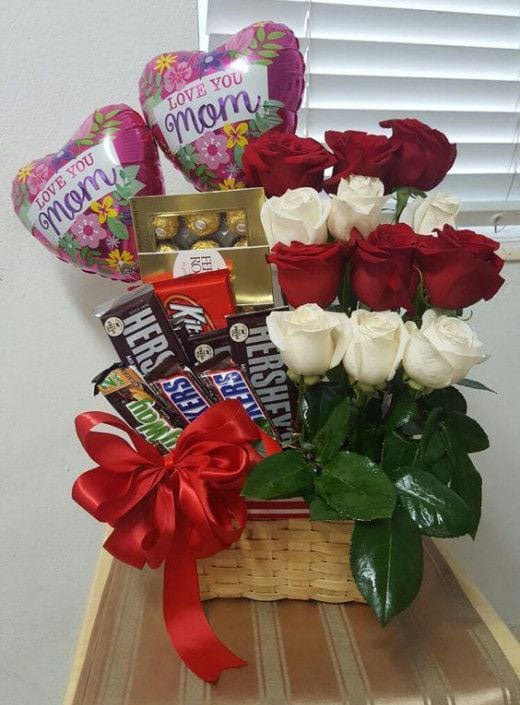 A gift basket containing red and white roses, chocolate candies, and heart-shaped balloons with 'I love you mom' printed on them.