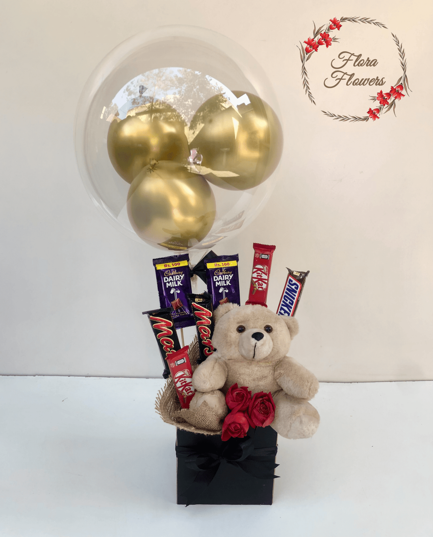 A gift basket containing chocolates, balloons, a teddy bear, and roses presented in a black box with a gold bow.