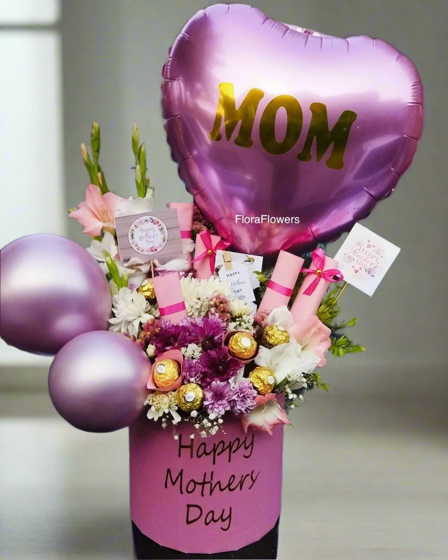 A gift arrangement featuring a pink heart-shaped balloon with the word 'MOM' written in gold, various pink and white flowers, green foliage, and decorative elements, presented in front of a pink box with 'Happy Mother's Day' written on it.