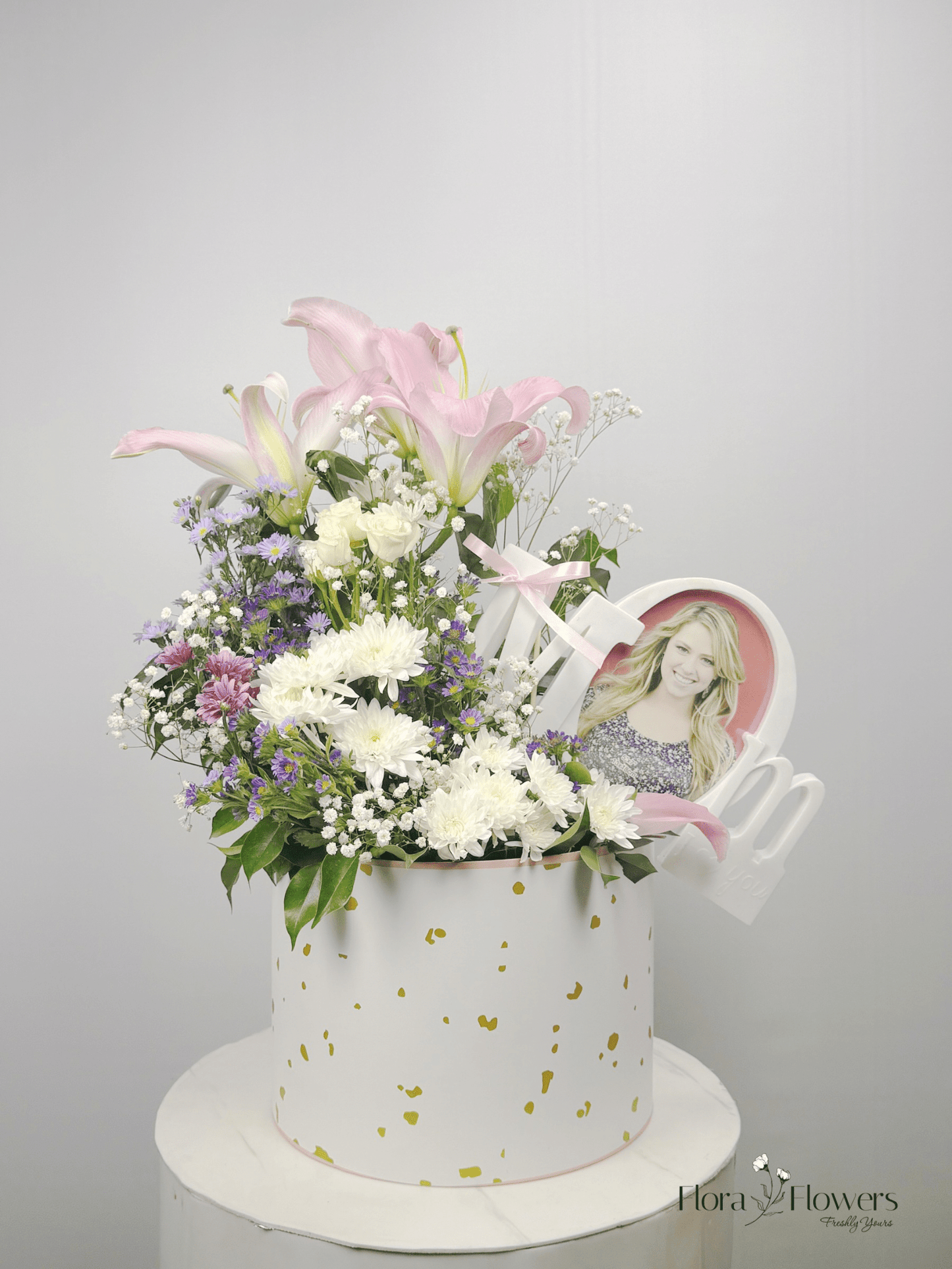 A basket arrangement featuring a lily, spray roses, baby's breath, and chrysanthemums in white, pink, and purple colors, with a photo frame added as a decorative element. Send flowers to islamabad. send flowers to pakistan