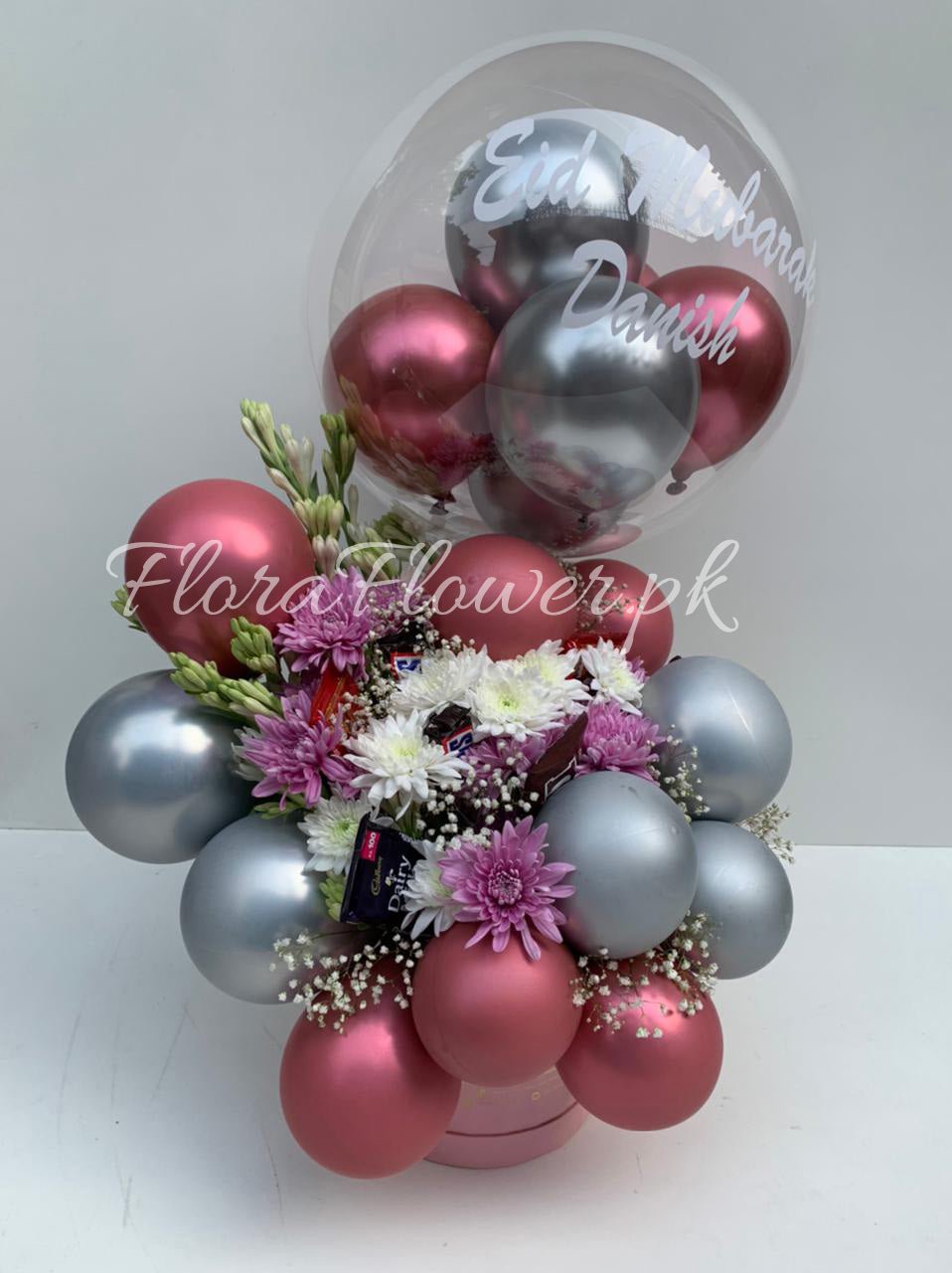 A bunch of balloons with one large balloon in the center that has text written on it, surrounded by smaller balloons in shades of pink, red, and silver, and adorned with imported chrysanthemums and baby's breath.