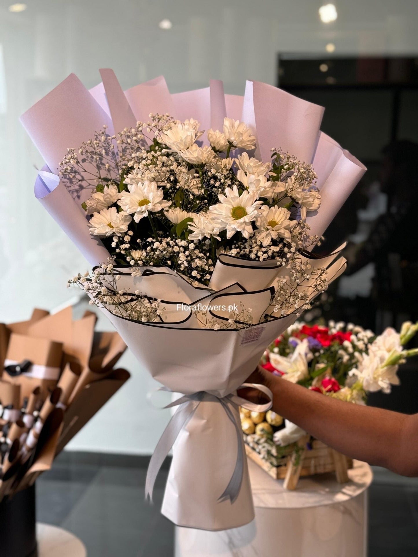 Bouquet of flowers wrapped in white paper with a pink ribbon, set against a blurred indoor background.