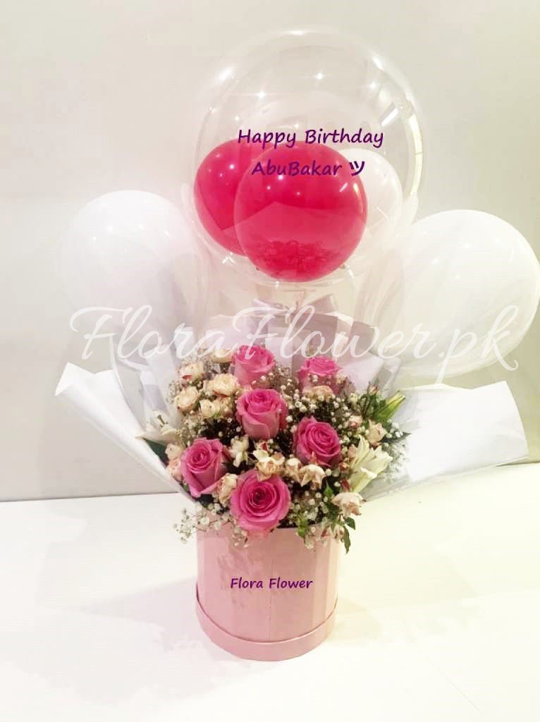 A gift flower basket containing an arrangement of pink roses, with two red balloons and decorative elements, presented in a clear cylindrical vase with a 'Happy Birthday' message on one of the balloons.