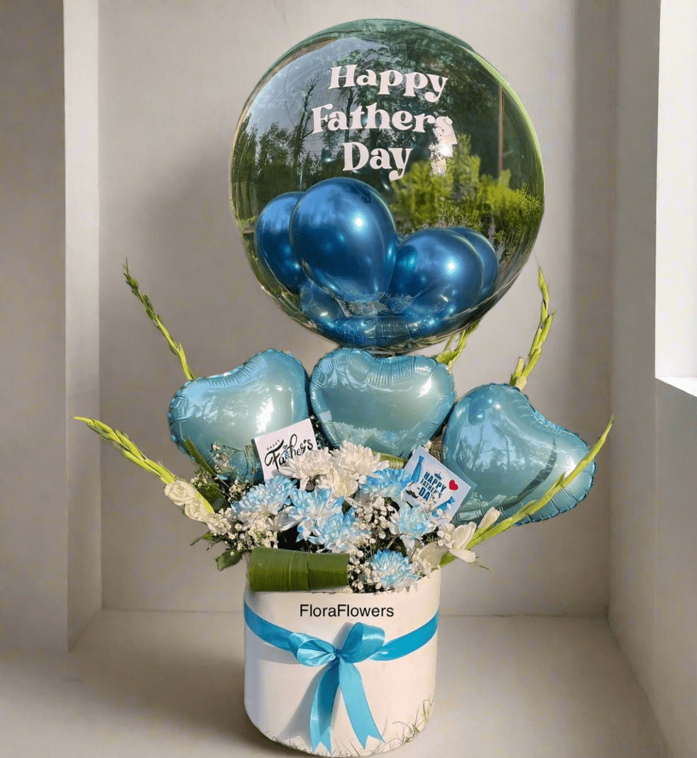 A bouquet of blue flowers with a transparent balloon carrying a 'Happy Father's Day' message, presented in a round white basket with a blue ribbon.