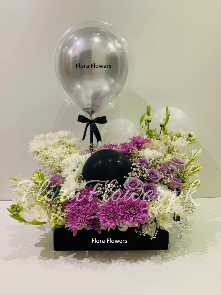 A floral arrangement featuring purple and white flowers with greenery, accompanied by balloons, in a black European tray with the brand name 'Flora Flowers' visible.