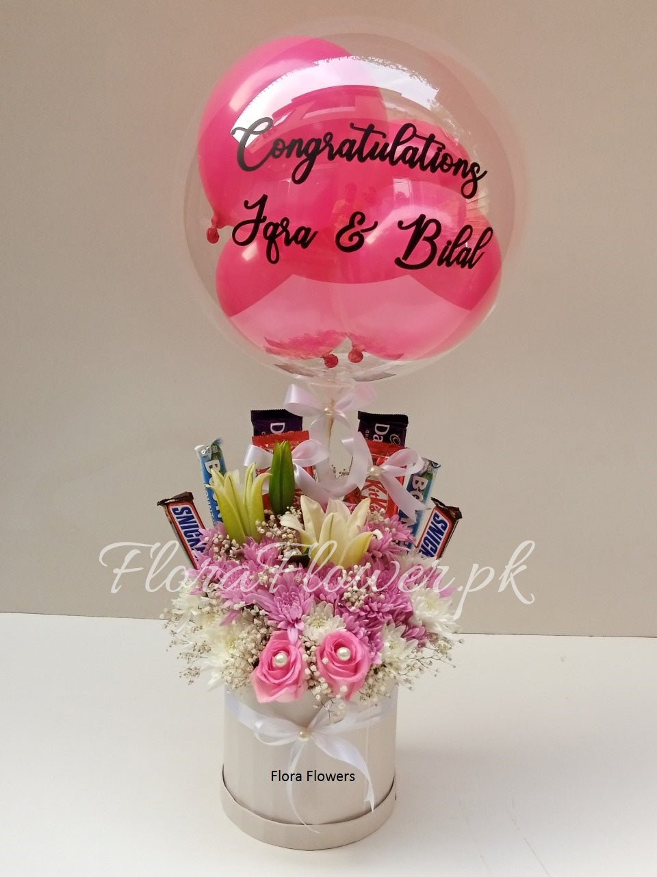 A gift arrangement featuring a pink balloon with text, multiple imported roses, chrysanthemums, baby's breath, a lily, and an assortment of chocolates.