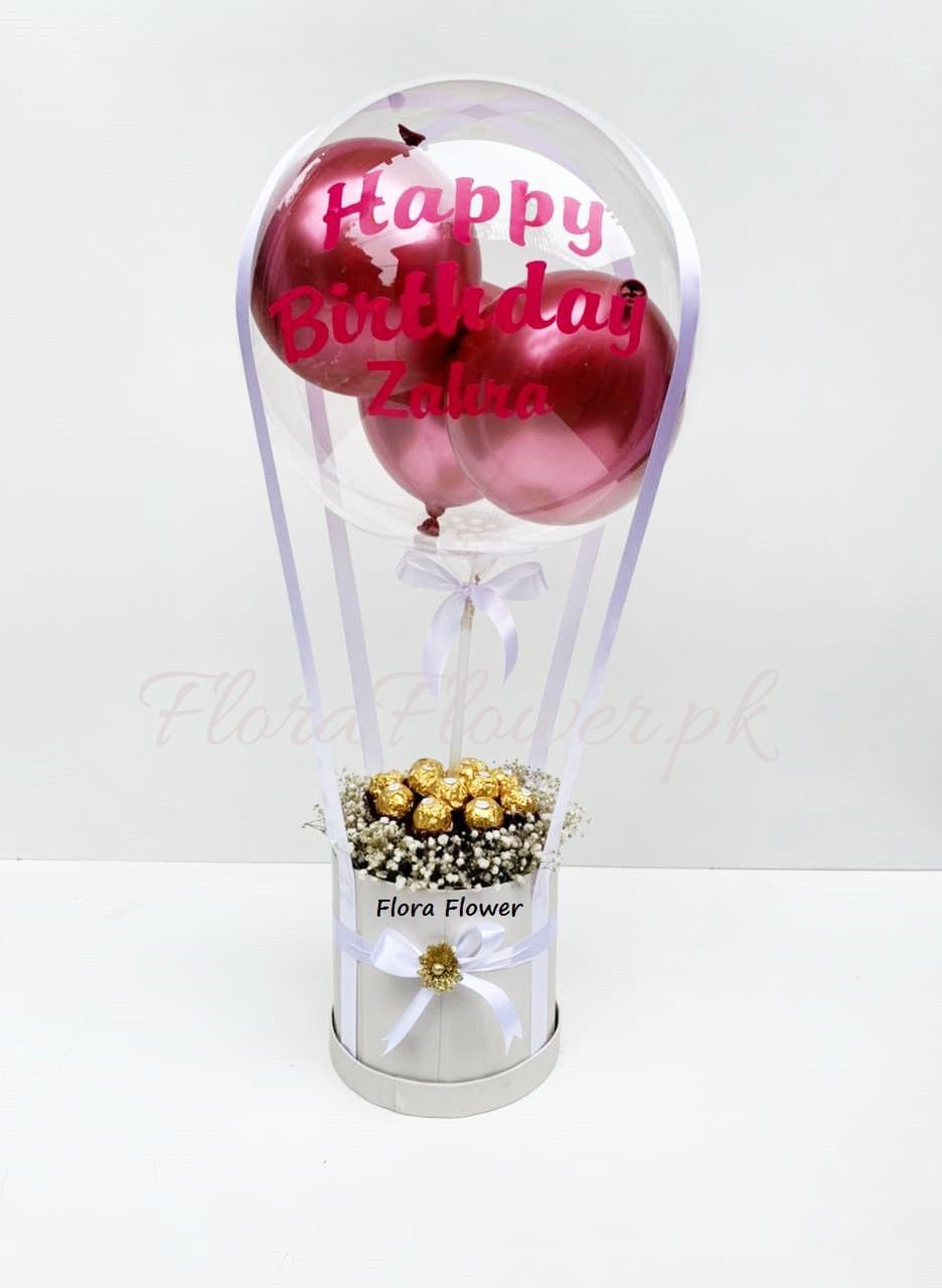 A gift basket containing a transparent balloon with pink text reading 'Happy Birthday', wrapped with a ribbon, alongside Ferrero Rocher chocolates and Baby's Breath flowers.