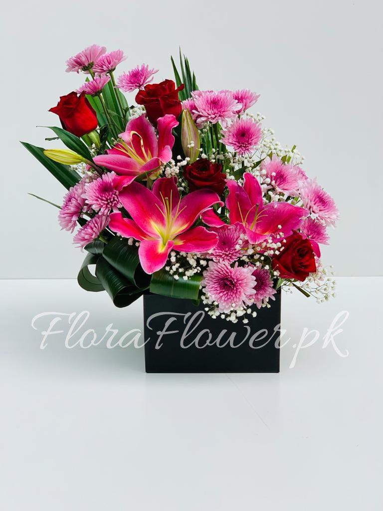 A floral arrangement featuring purple chrysanthemums, red roses, and lilies with green leaves in a black container, with the watermark 'FloraFlower.pk' visible.