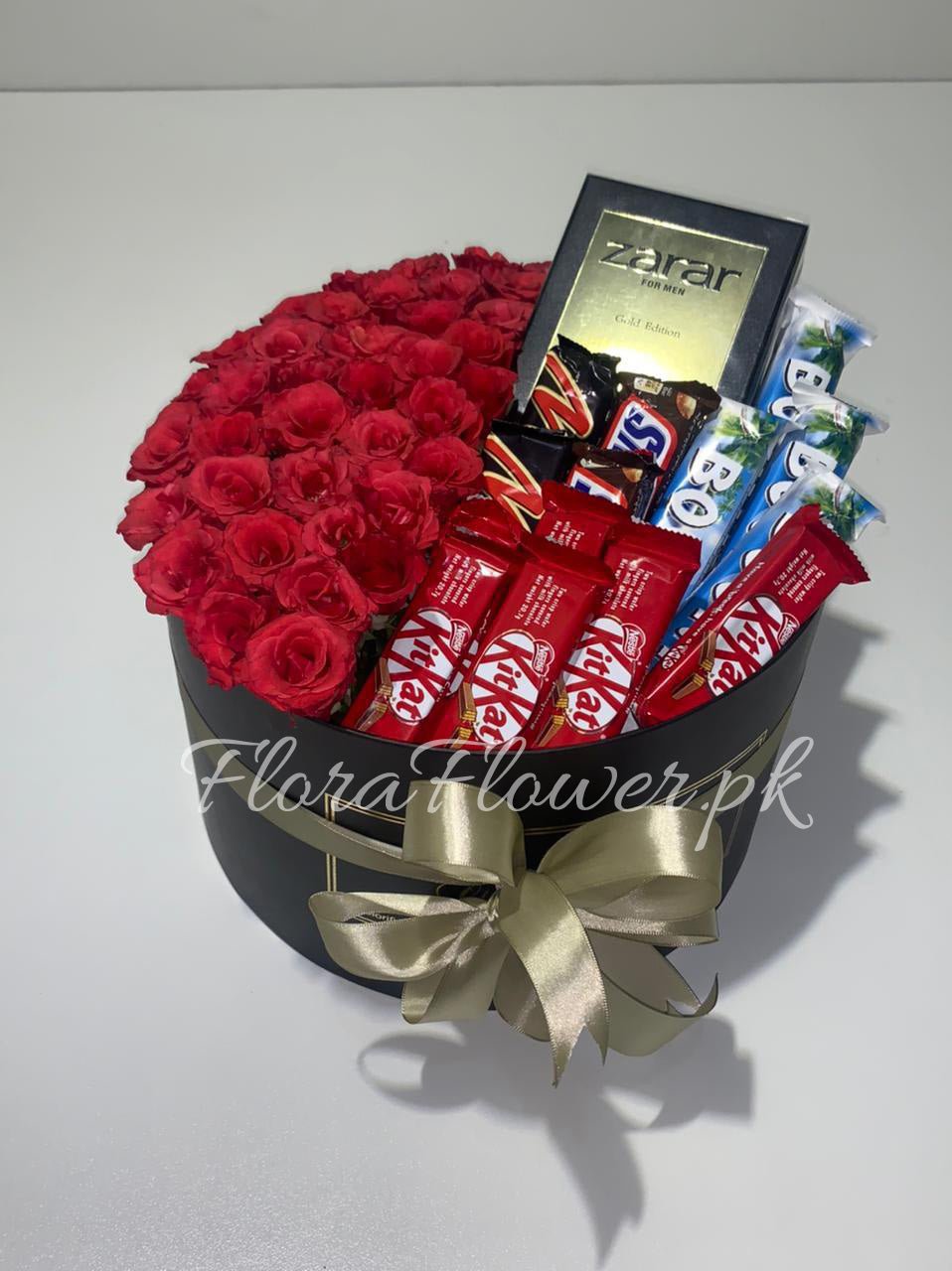 A round gift basket containing red roses, various chocolates, and a small bottle of perfume, adorned with a gold ribbon.