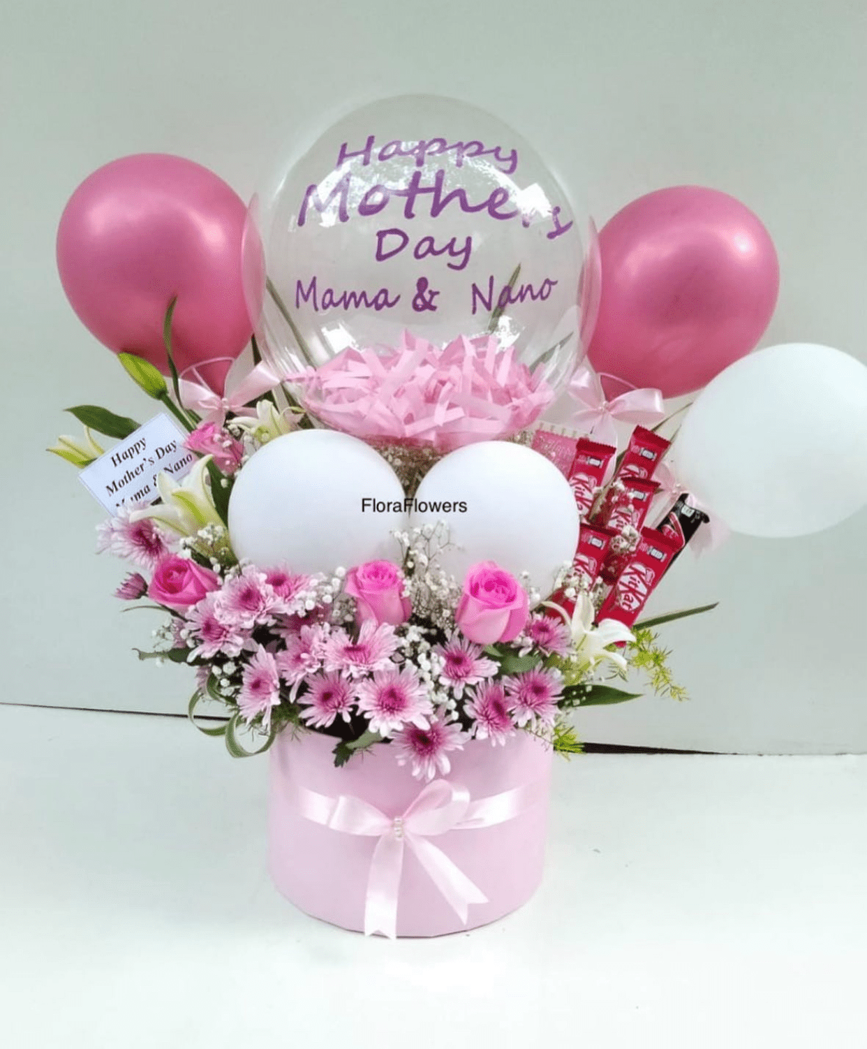 A gift arrangement featuring an assortment of pink and white flowers, chocolates, and balloons, with a large transparent balloon displaying text.