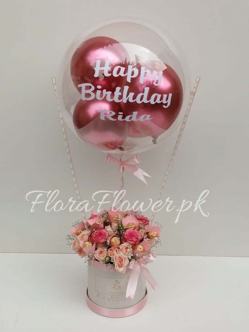 A pink balloon with 'Happy Birthday' text, attached to a pink and white floral arrangement in a round container.