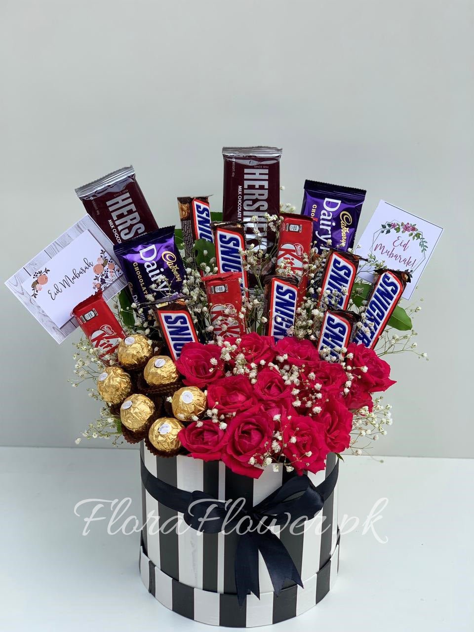 A gift basket arrangement consisting of red roses, baby's breath, and an assortment of chocolates including Ferrero Rocher and Snickers, presented in a black and white striped cylindrical container.