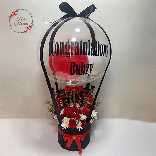 A balloon bouquet with a large clear balloon that reads 'Congratulations Bubzy', decorated with red and white flowers, chocolates, and a red balloon.