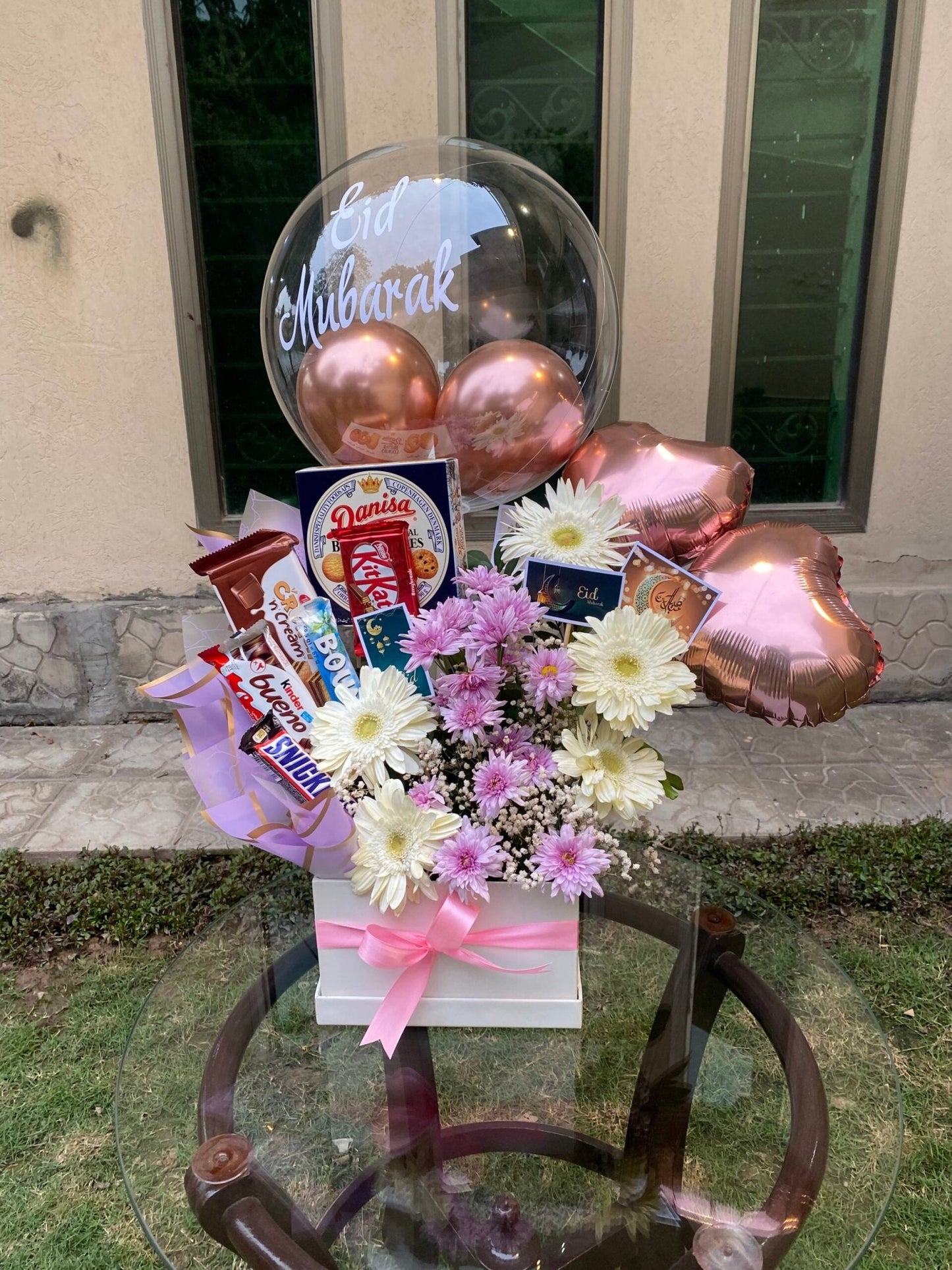 A customized balloon arrangement featuring a transparent balloon with printed text, accompanied by flowers, chocolates, and cookies displayed on a table.