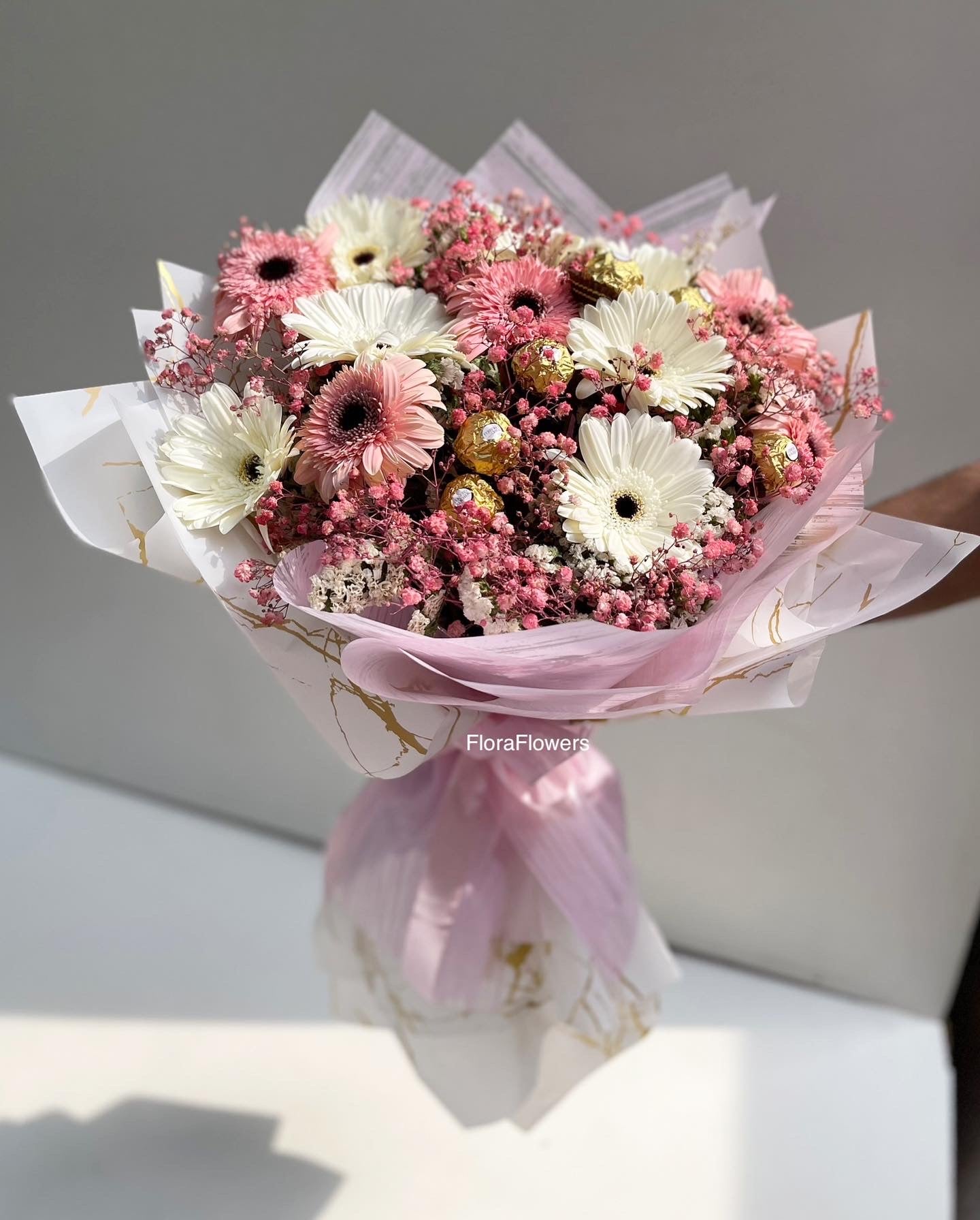 A bouquet of pink, white, and red flowers with green leaves, wrapped in pink paper with gold accents.