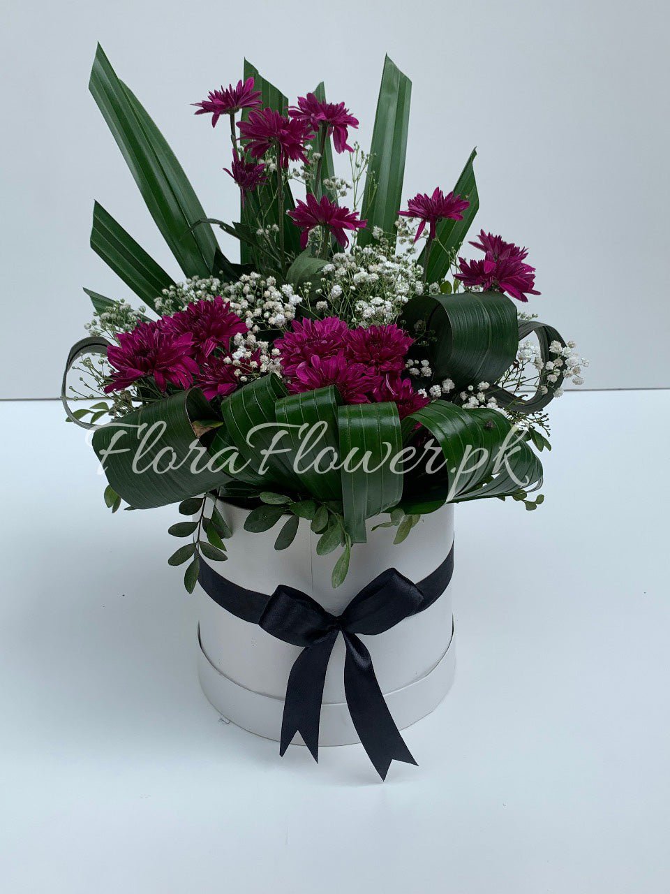 A floral arrangement featuring purple chrysanthemums, white baby's breath, and green leaves, presented in a round white container with a black ribbon.