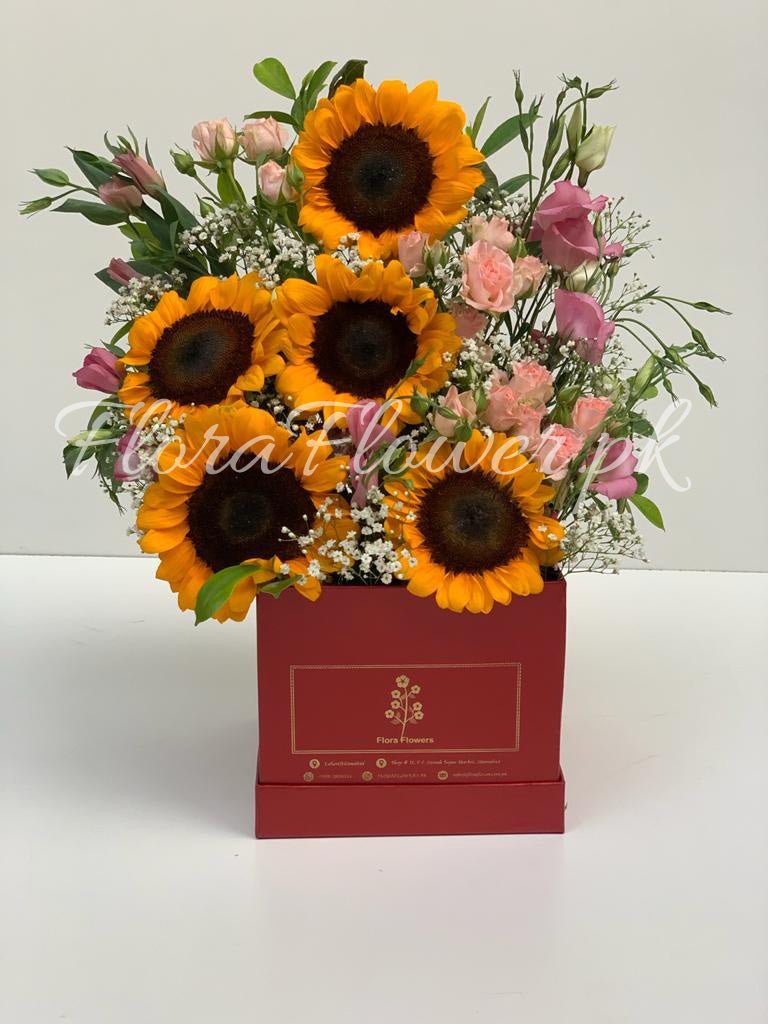 A bouquet of flowers including sunflowers, spray roses, and baby's breath presented in a red square-shaped vase with a floral design.