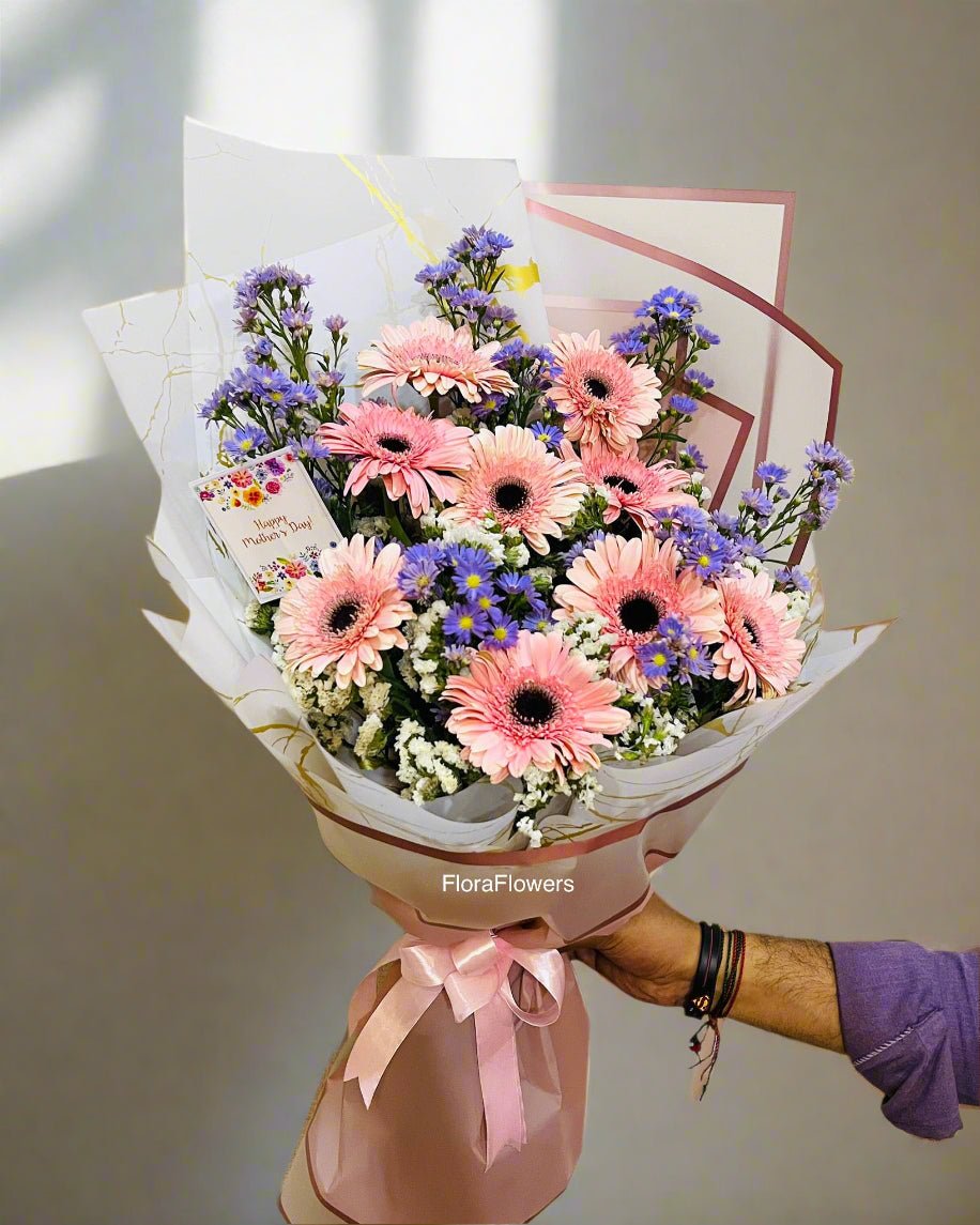 A bouquet of flowers including pink and purple gerberas, baby's breath, and other complementary foliage, wrapped in white paper with a pink ribbon.