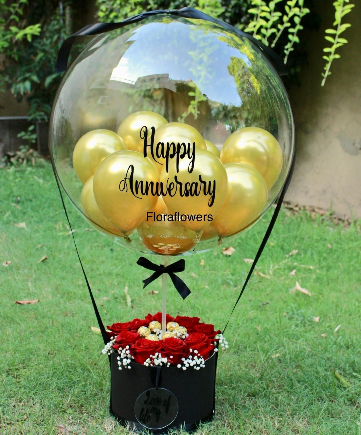 A gift basket with red and white flowers, topped with a transparent balloon that says 'Happy Anniversary' in gold lettering, presented on grass.