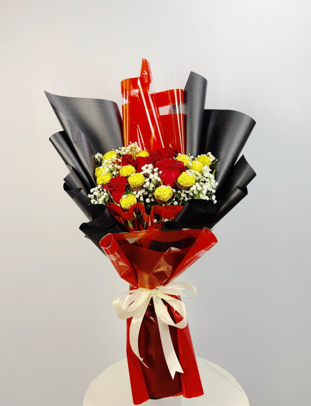 Bouquet of flowers wrapped in red and black paper with a white ribbon on a light gray background. Send flowers to lahore. send flowers to Pakistan