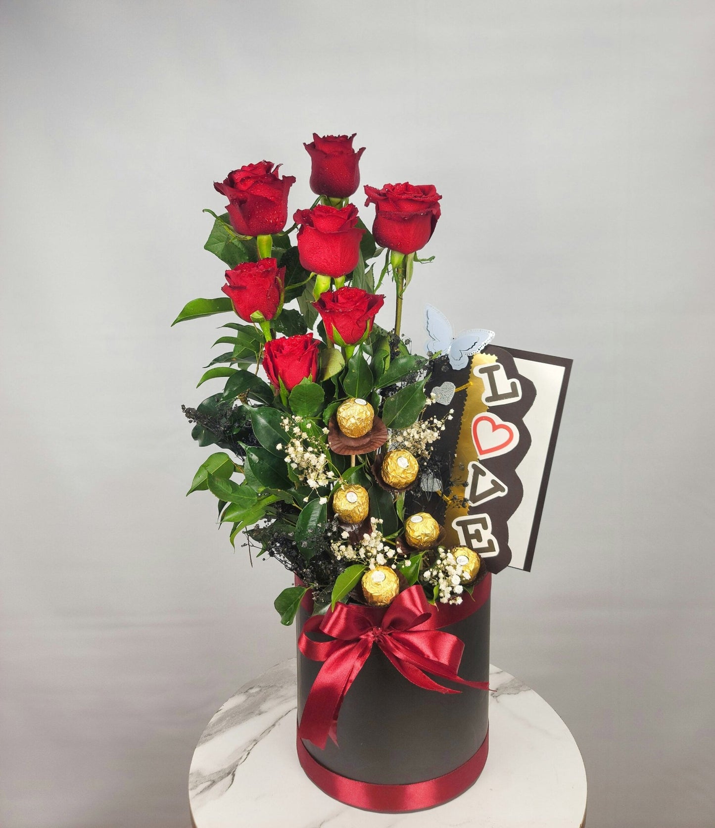 Bouquet of red roses in a black box with a red ribbon, surrounded by chocolates and a 'I love you' card on a light gray background. Send flowers to islamabad. send flowers to pakistan