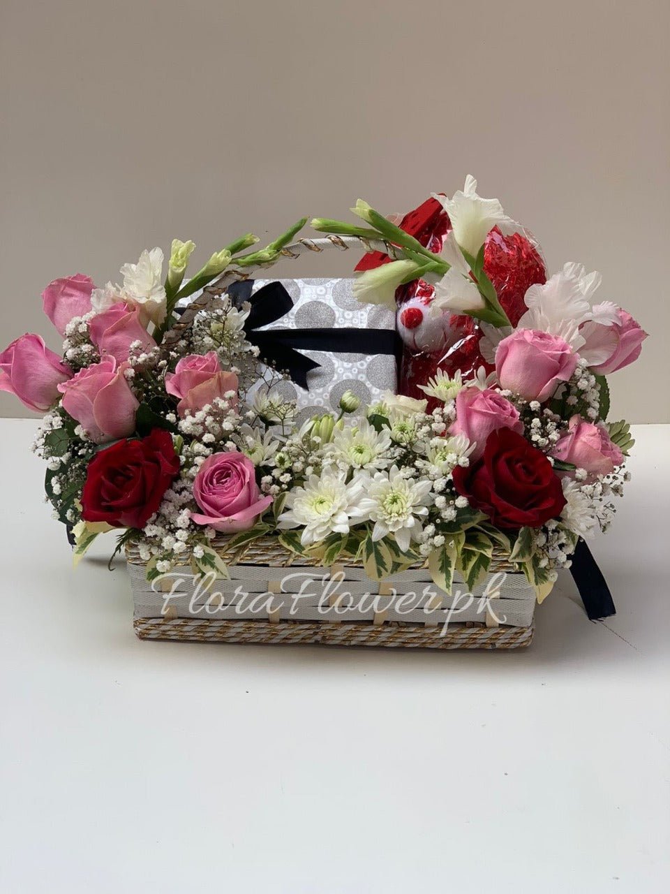 A basket arrangement of various flowers including red and pink roses, white chrysanthemums, and baby's breath with a box of chocolate.