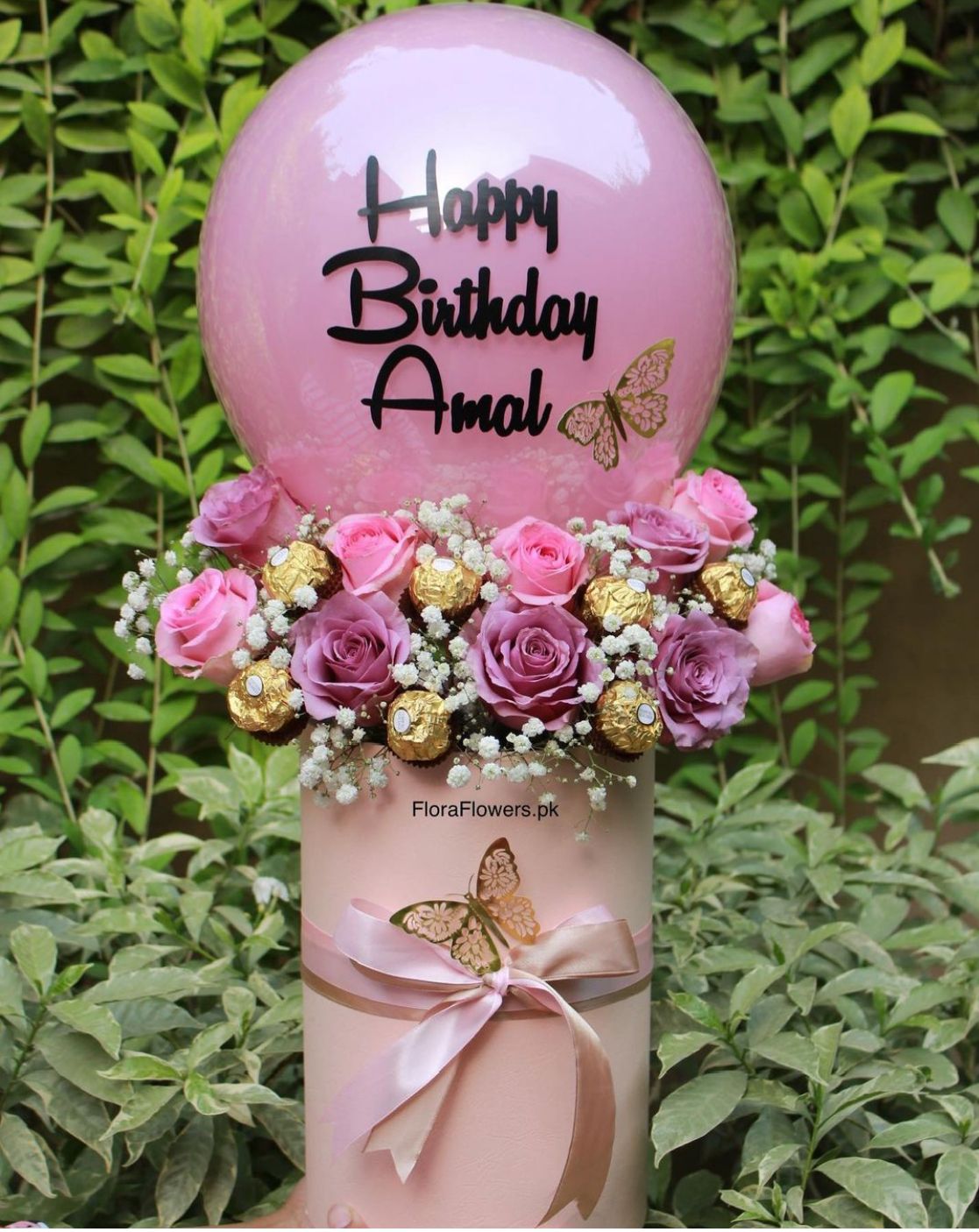 A balloon bouquet arrangement with a pink balloon reading 'Happy Birthday Amal' decorated with pink and purple roses, Ferrero Rocher chocolates, and baby's breath, presented on a table.