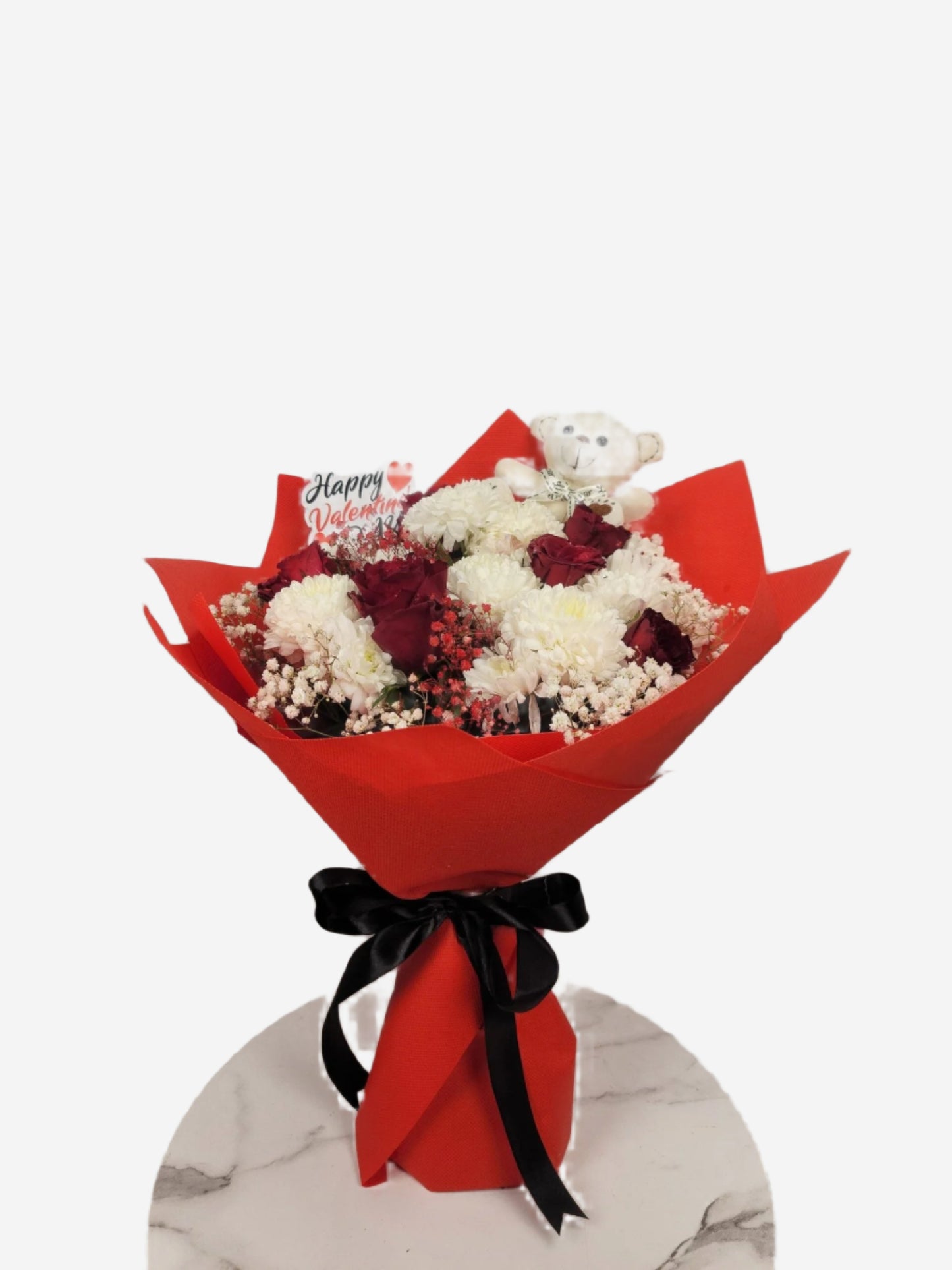 Bouquet of flowers wrapped in red paper with a teddy bear and 'Happy Valentine's Day' card on a white marble surface.