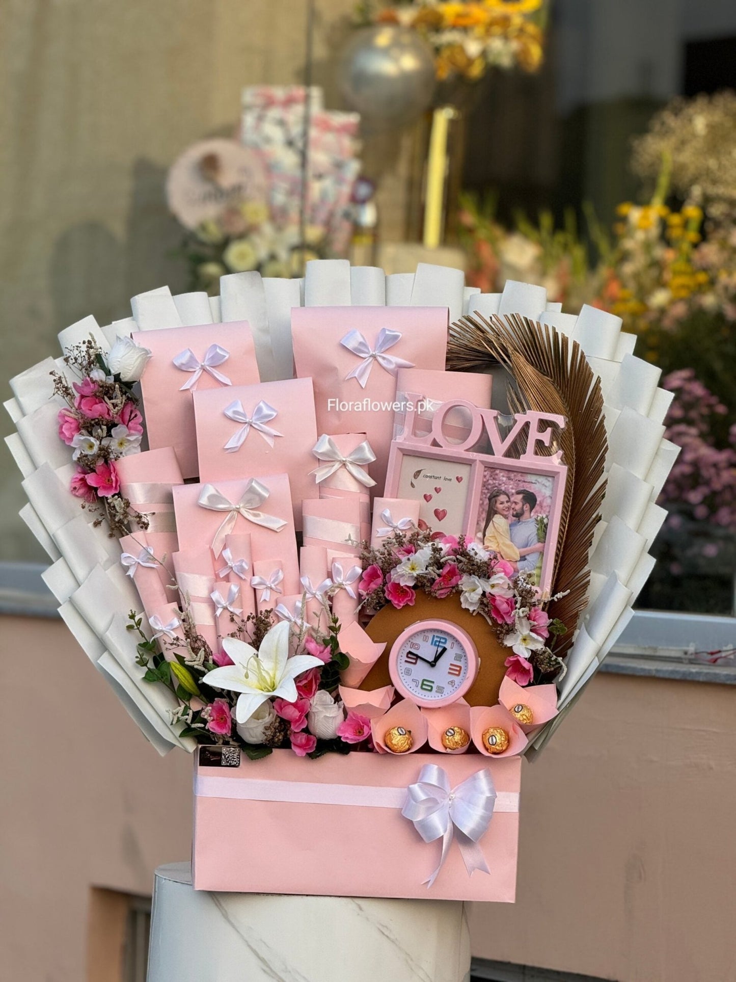 Bouquet with pink elements, including a clock and photo frame, against a blurred floral background.