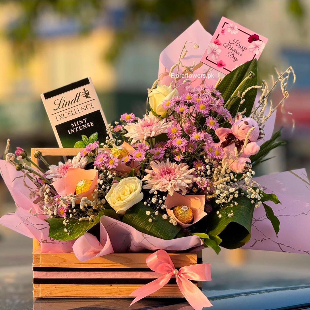 A gift basket containing an assortment of flowers, chocolates, and a teddy bear arranged in a wooden basket with a pink bow.
