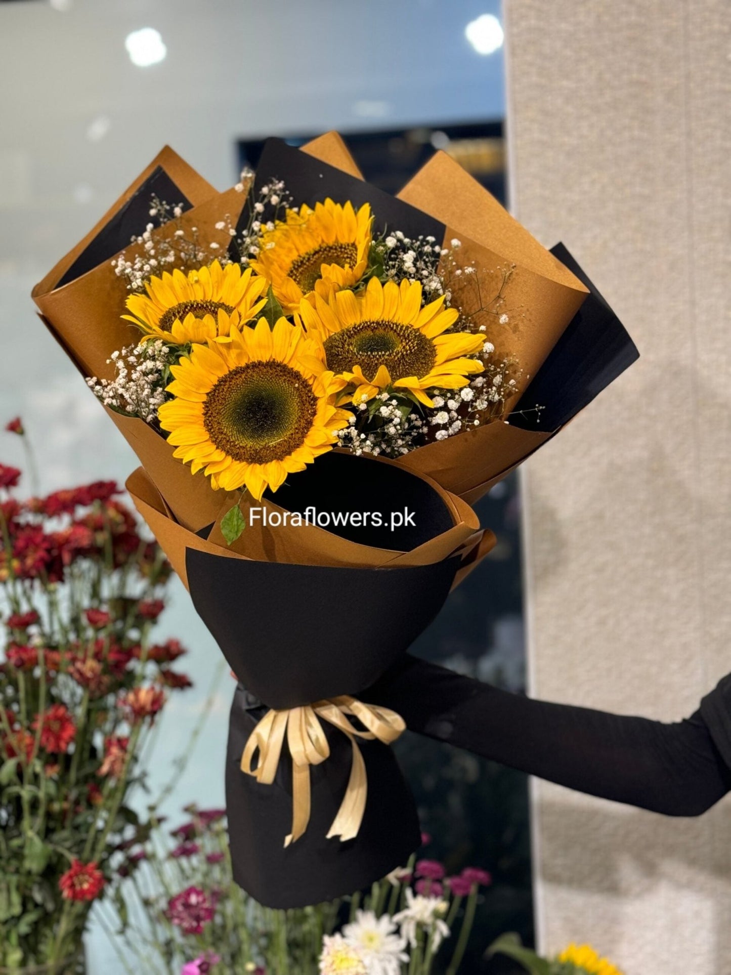 Bouquet of sunflowers wrapped in black and brown paper with a gold ribbon, held by a person. Brand 'Floraflowers.pk' visible.