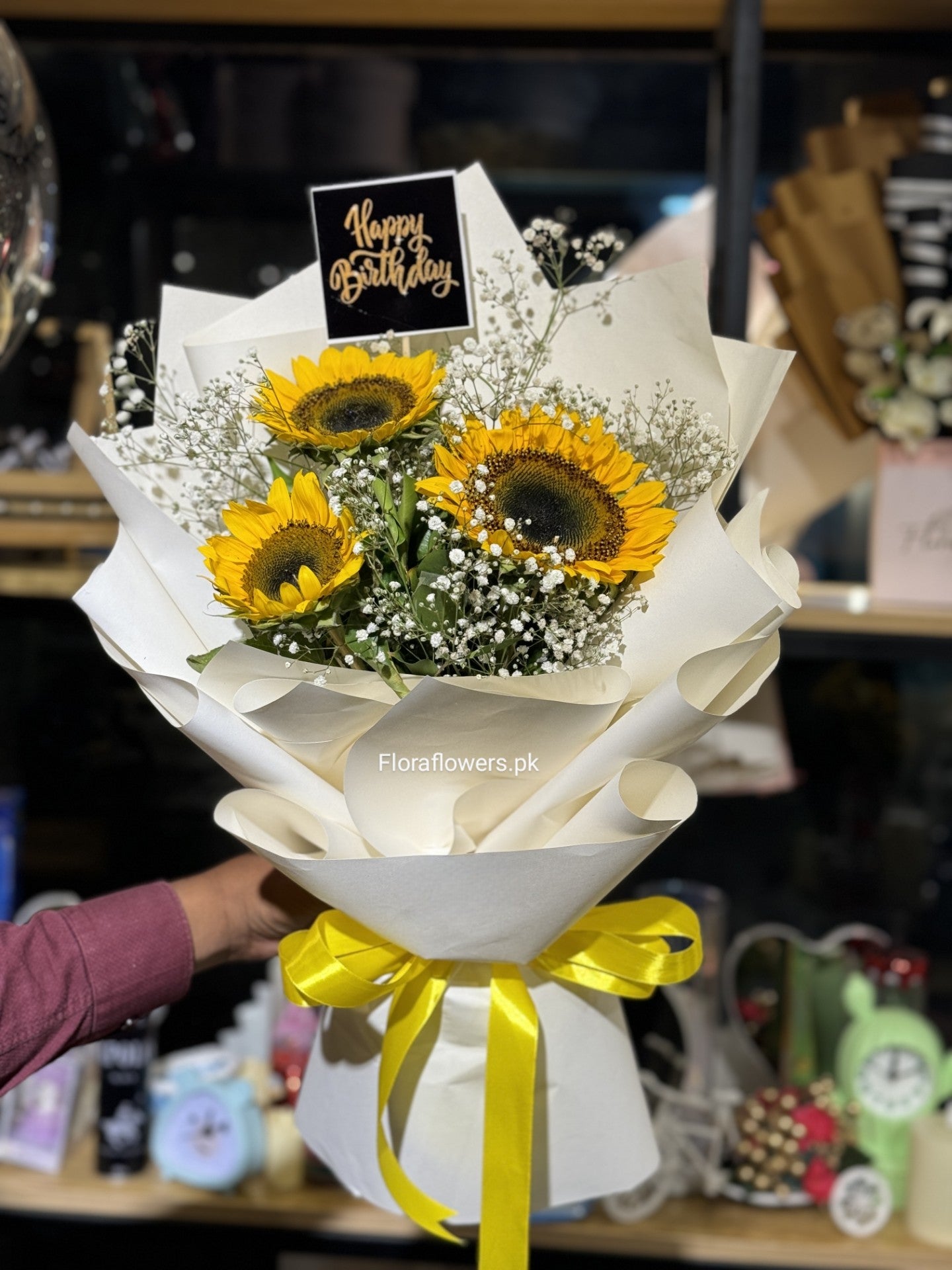 A bouquet of flowers consisting of sunflowers and Baby's Breath, wrapped in white paper with a yellow ribbon, presented by Flowersonline.uk.