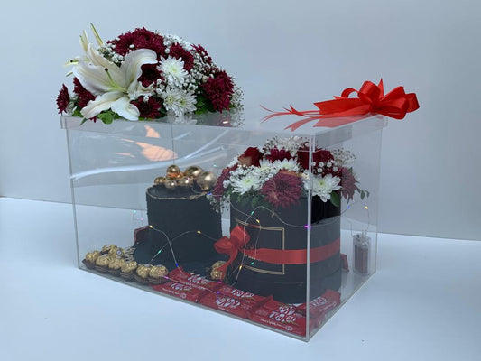 An acrylic gift box containing a basket of red, white, and purple flowers, assorted chocolates, and decorative lights.