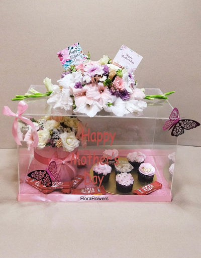 A gift box containing a bouquet of flowers, cupcakes, and other Mother's Day related items, adorned with decorative elements in pink tones.