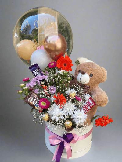 A teddy bear embracing a round transparent balloon, with an assortment of flowers, balloons, and chocolates arranged in a box. Send flowers to islamabad. send flowers to pakistan