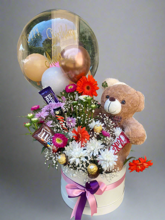 A teddy bear embracing a round transparent balloon, with an assortment of flowers, balloons, and chocolates arranged in a box.