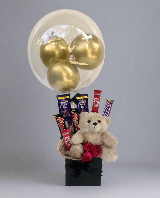 A gift basket containing chocolates, a teddy bear, and balloons with a clear balloon on top, arranged with red roses.