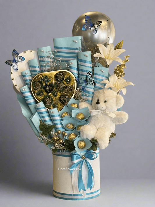 A bouquet arrangement composed of various chocolates including Snickers, Mars, KitKat, Dairy Milk, a wafer, and Ferrero Rocher, adorned with blue ribbons and decorative elements, and accompanied by a small teddy bear.