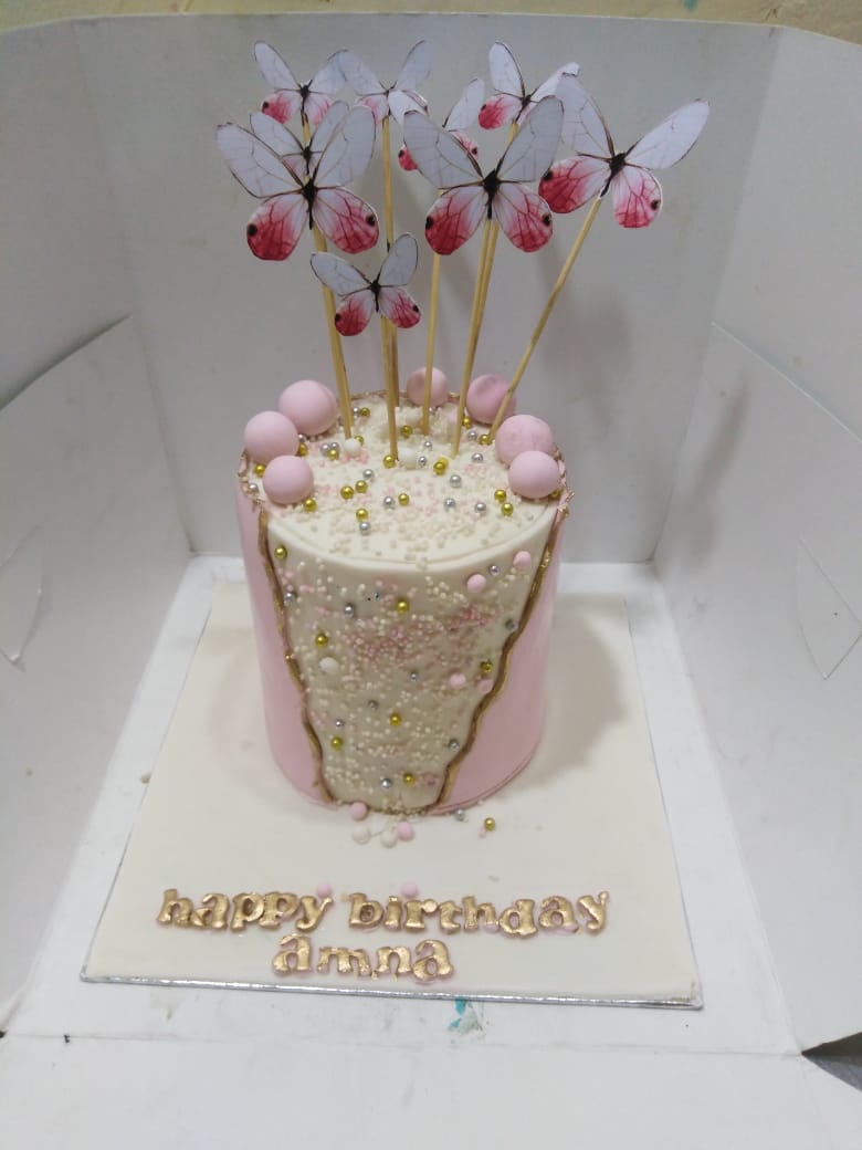 Decorative cake with butterfly toppers on a white stand, featuring the text 'happy birthday amna'.