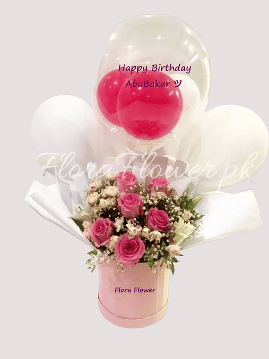 A gift basket arrangement featuring a round balloon with 'Happy Birthday' text, a vase with pink roses, spray roses, a lily, and baby's breath, all presented by Flora Flower.