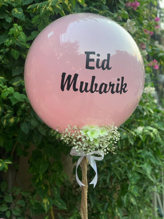 Pink balloon with 'Eid Mubarak' text and floral decorations against a green leafy background