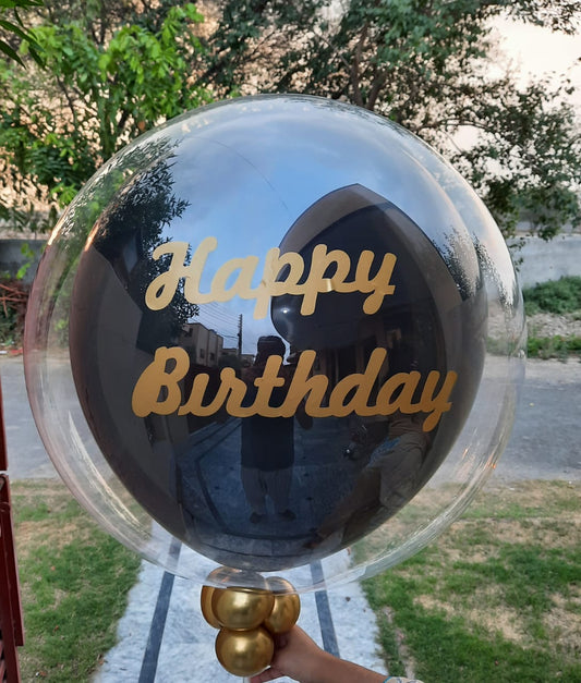 Transparent balloon with 'Happy Birthday' text and gold base held outdoors.