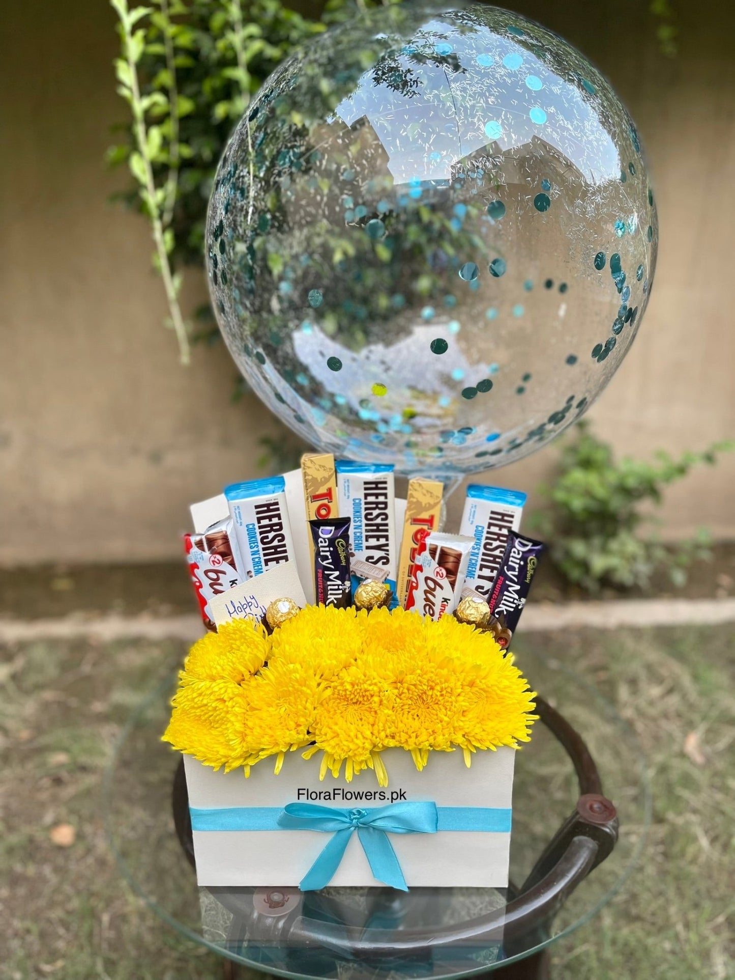 Gift basket with candy bars and yellow flowers on a small table outdoors. Send flowers to islamabad. send flowers to pakistan