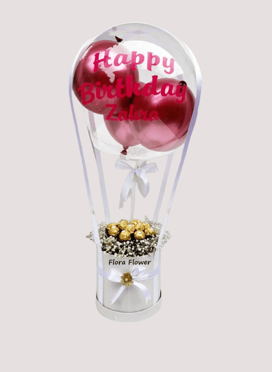 A gift basket containing Ferrero Rocher chocolates and Baby's Breath flowers, presented in a clear balloon-shaped container with a 'Happy Birthday' message on a pink balloon, and decorated with a gold ribbon.