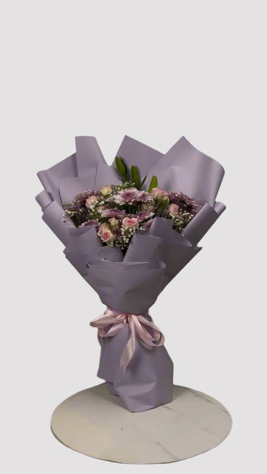 A bouquet of flowers including chrysanthemums, spray roses, a lily, and baby's breath, wrapped in purple paper with a pink ribbon.
